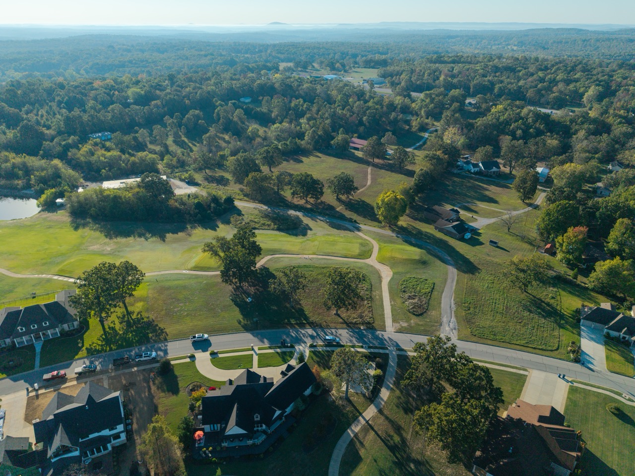 Aerial view of a suburban neighborhood adjacent to a golf course, with winding cart paths, green fairways, scattered houses, a small pond, and dense trees extending to the distant forest.